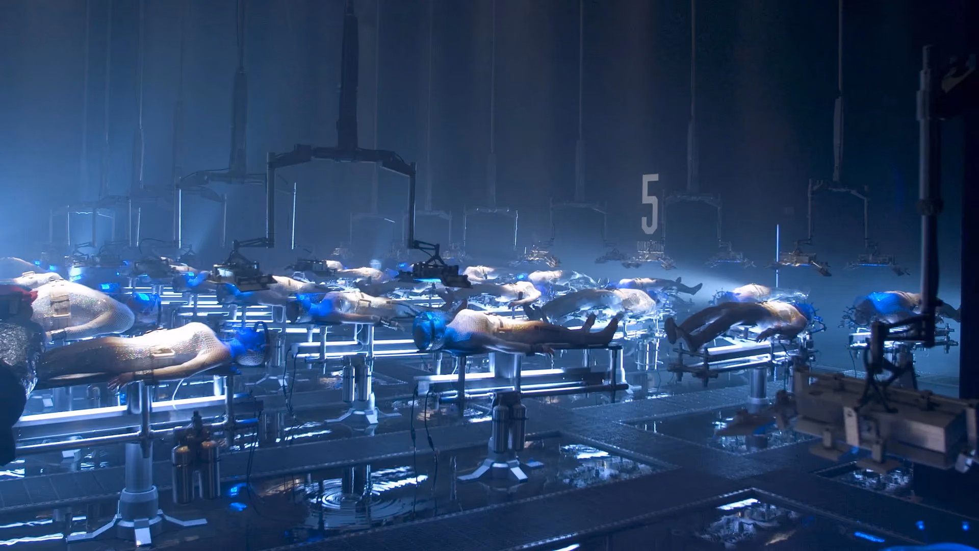 Rows of human bodies laid out on lit tables in a dark, blue-lit industrial hall