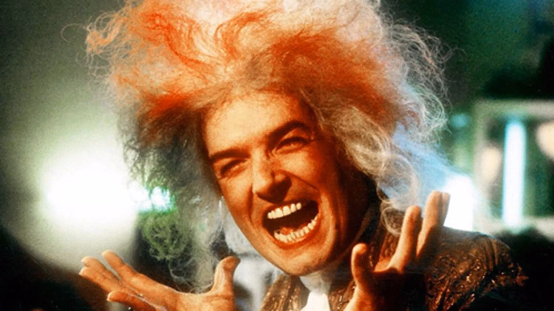 A man with wild orange hair laughing manically, both hands raised